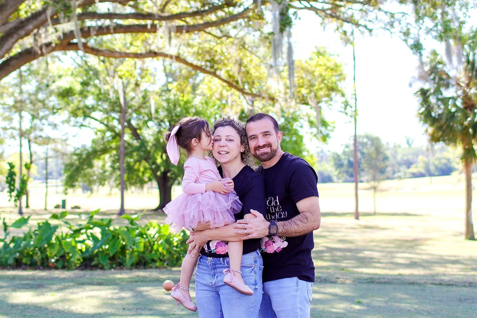 A family of three outdoors on a sunny day in a park, with a young girl giving a kiss on her mother's cheek, while her father smiles. The mother is holding the girl, and all are smiling, surrounded by trees and greenery.