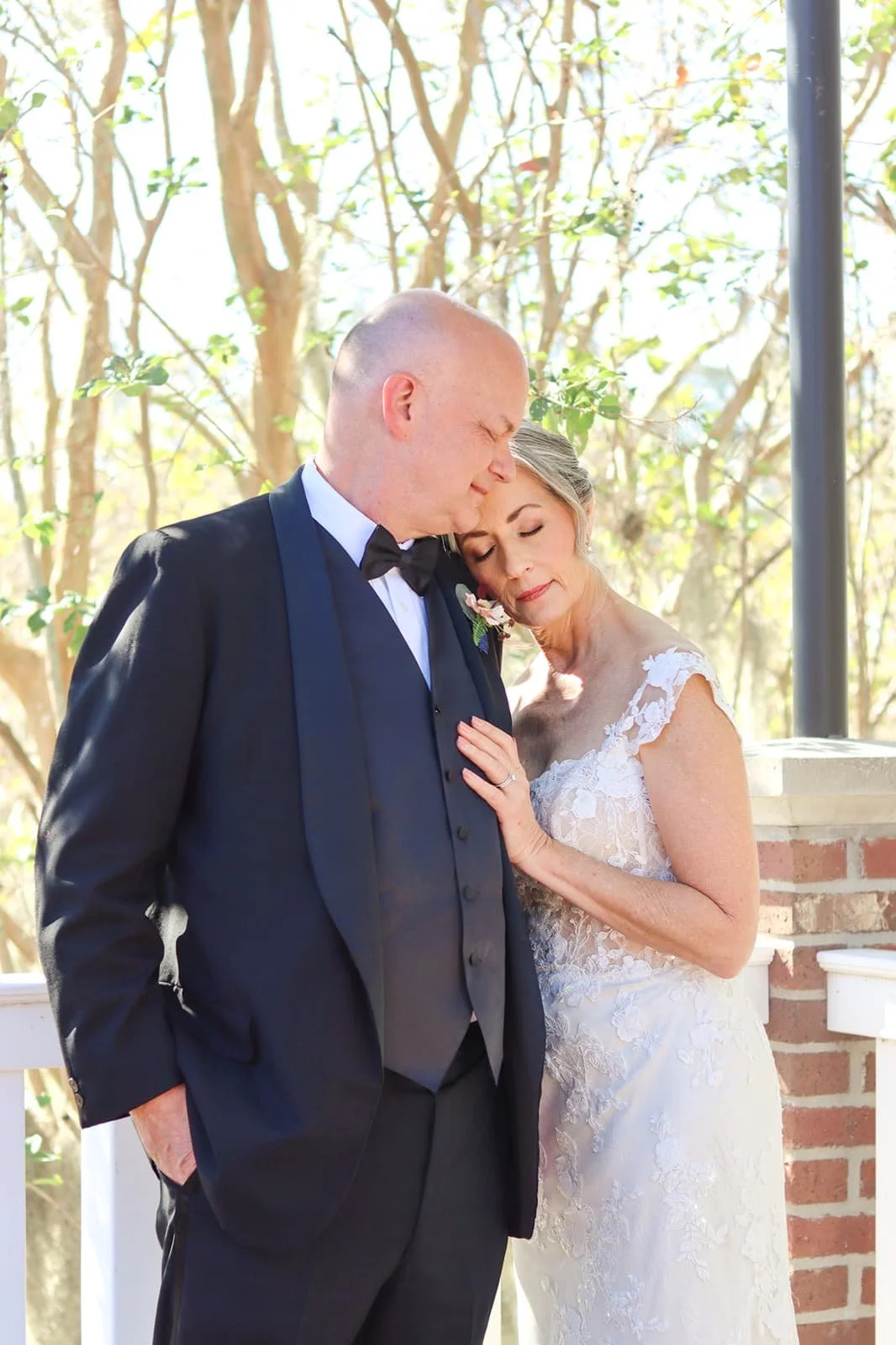 A bride and groom sharing a tender moment outdoors, with the bride leaning her head on the groom's forehead. The groom is dressed in a black tuxedo with a bow tie, while the bride wears a white lace wedding gown. They are surrounded by trees and sunl