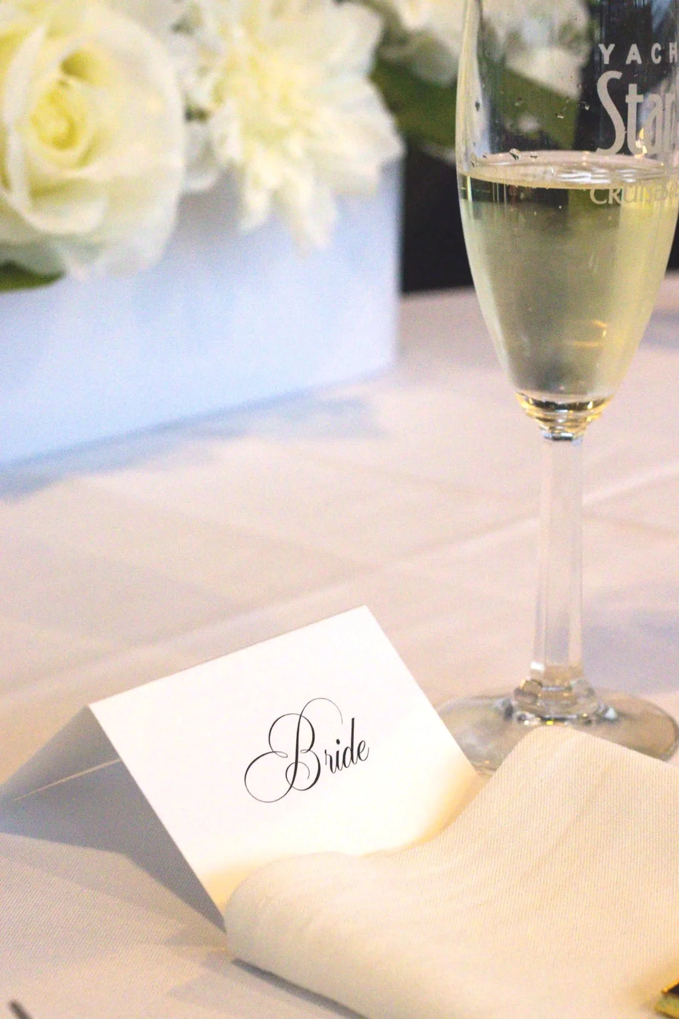 A white place card with the word 'Bride' written in elegant black script, placed on a table with a white napkin, next to a glass of champagne, and a vase of white flowers in the background.