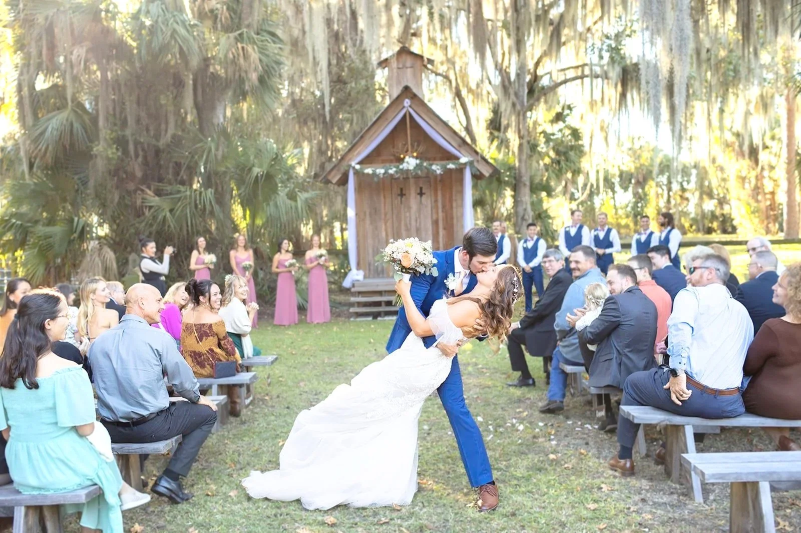 A bride and groom are dancing and kissing at an outdoor wedding ceremony. The bride is wearing a white wedding dress and the groom is in a blue suit. Guests are seated on benches on either side, with some clapping and smiling. Wooded trees and a smal
