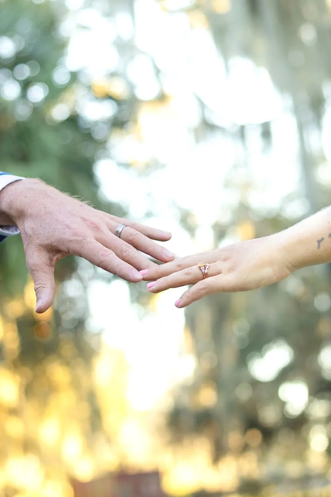 Close-up of two hands reaching toward each other outdoors, with a blurred green and yellow background, one hand has a wedding ring and the other has a tattoo.