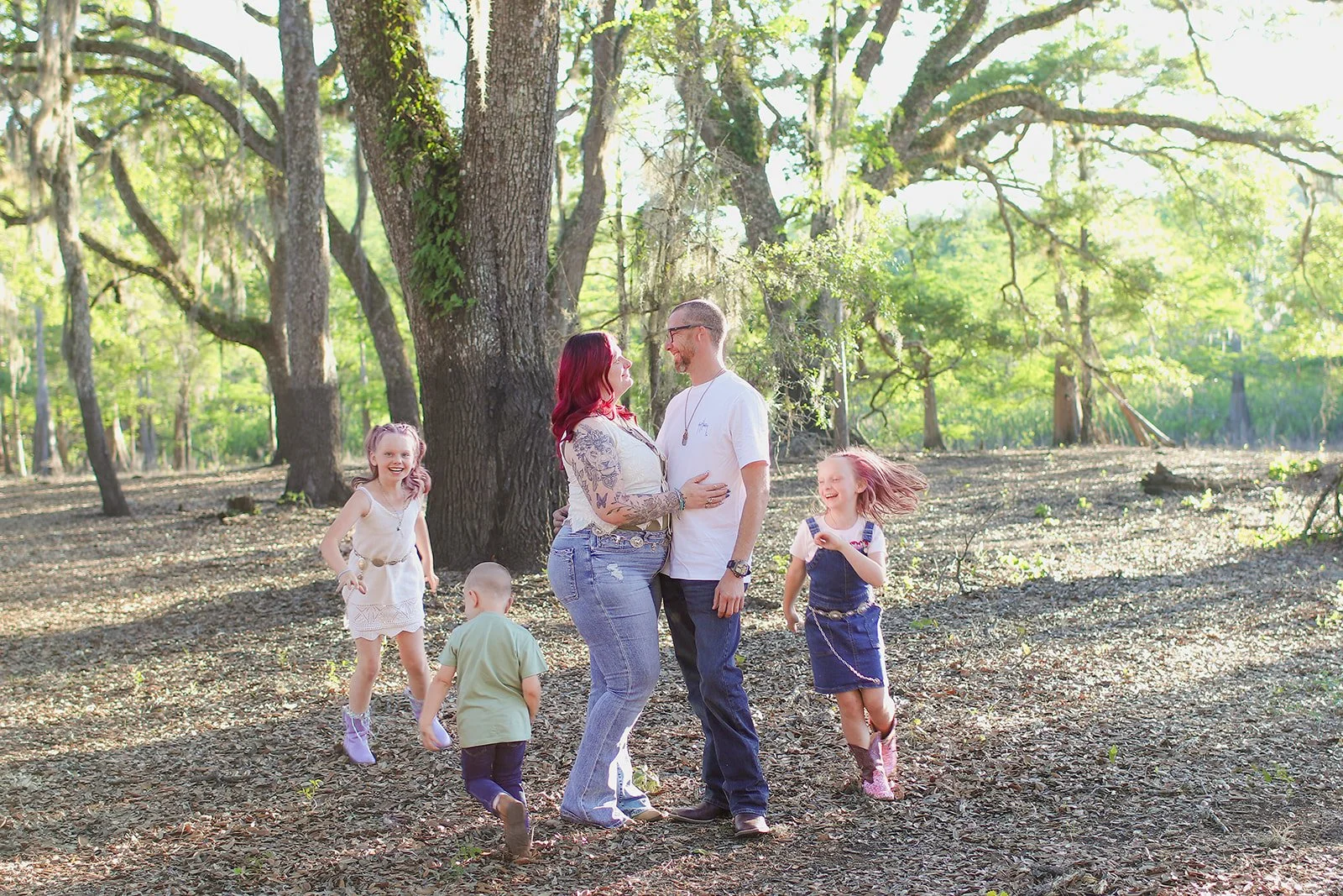 A family of five, including a woman with red hair, a man, and three young children, playing and smiling in a wooded park with tall trees and sunlight filtering through the leaves.