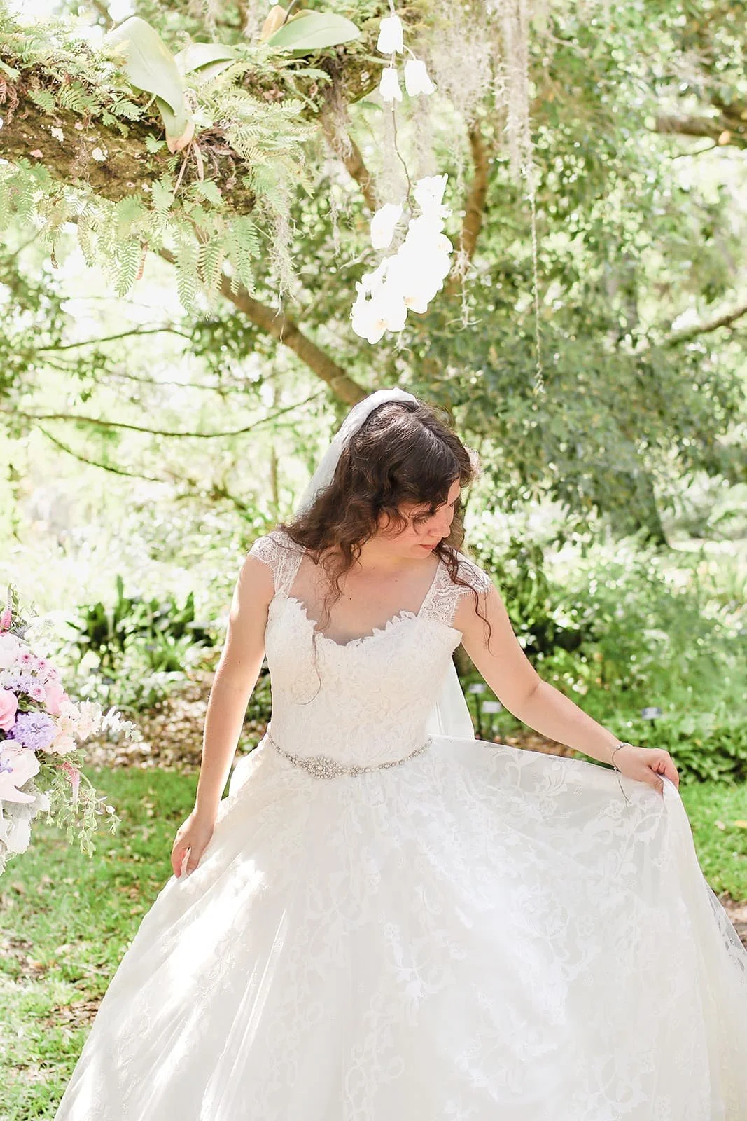 A bride in a white wedding dress with lace details and a beaded belt, holding the skirt and standing outdoors on grass, surrounded by greenery and hanging white flowers.