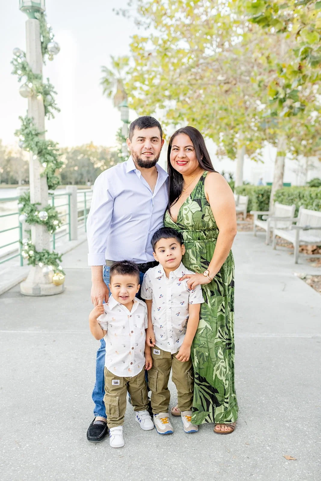 A family of four standing outdoors on a paved path near a body of water, with trees and benches in the background. The father and mother are standing behind two young boys, all smiling at the camera. The mother is wearing a green leafy dress, the fat