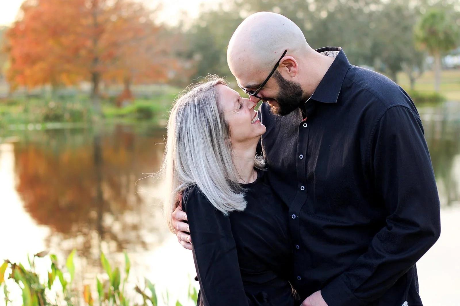 A couple embracing with their foreheads touching outdoors in front of a pond and autumn trees.