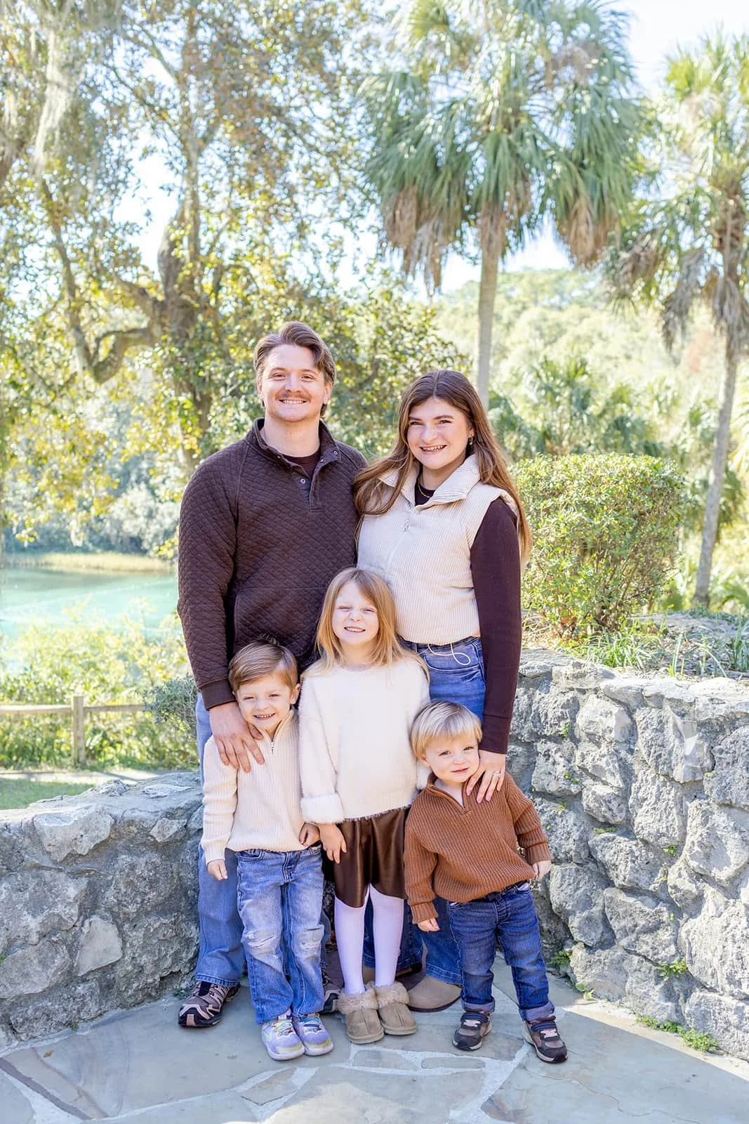 A family of six. Two adults and four children, standing outdoors near a stone wall with trees and a lake in the background. It's a sunny day.