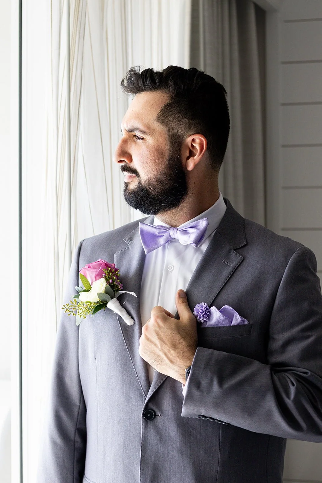 A man in a gray suit with a lavender bow tie and pocket square, holding his chest near his heart, standing by a window and looking outside.