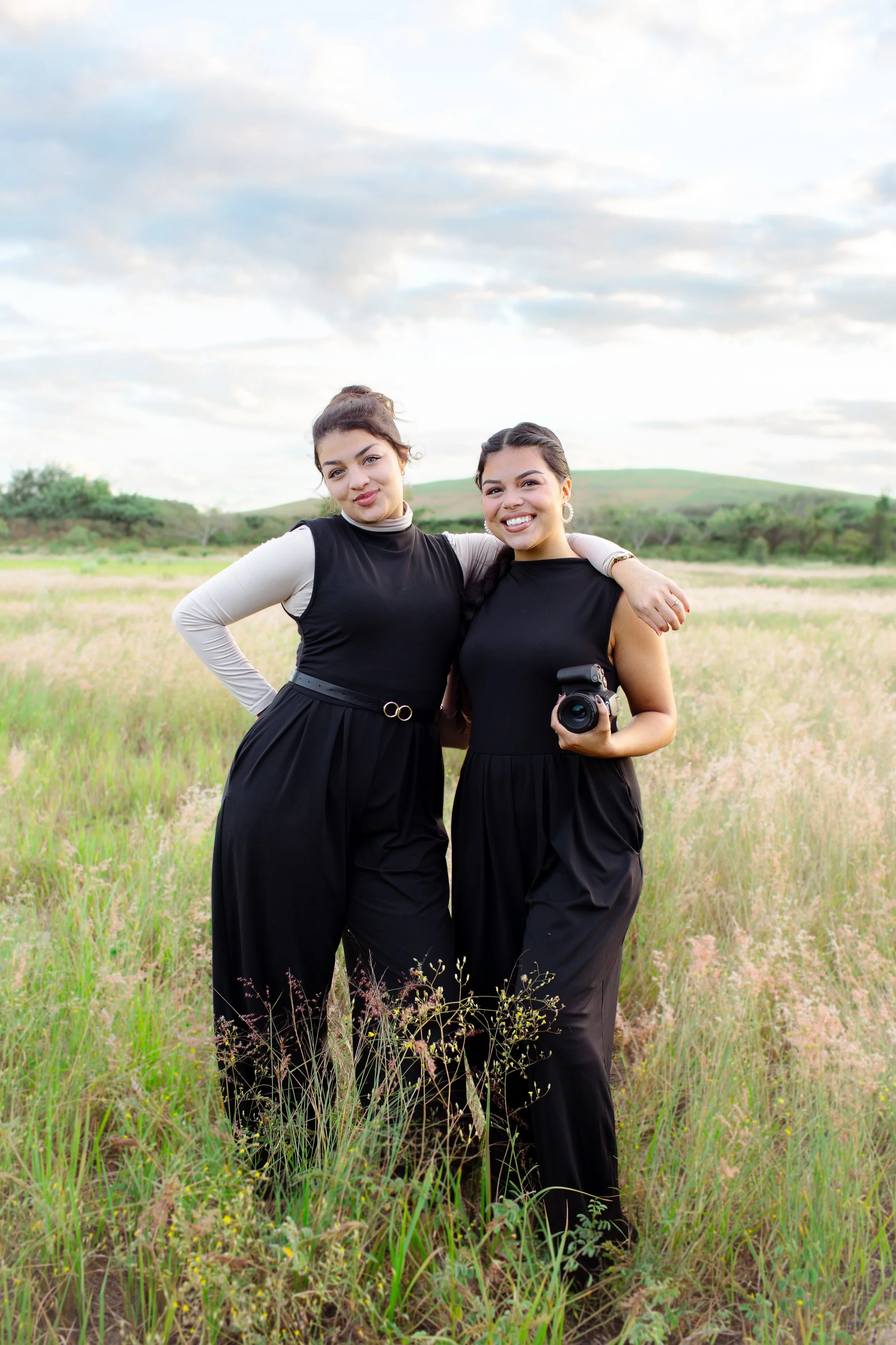 Sisters and lead wedding and family photographers Amaya and Krysta posed with cameras in field, smiling