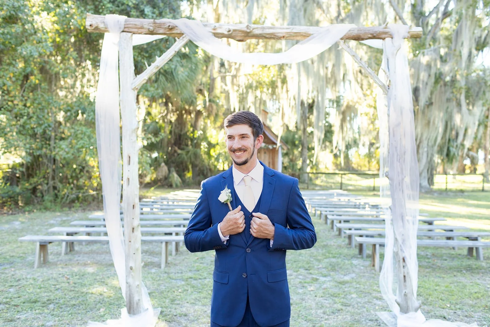 A man in a blue suit, white shirt, and beige tie standing outdoors under a wooden arch decorated with white fabric, smiling and holding a white flower boutonniere.