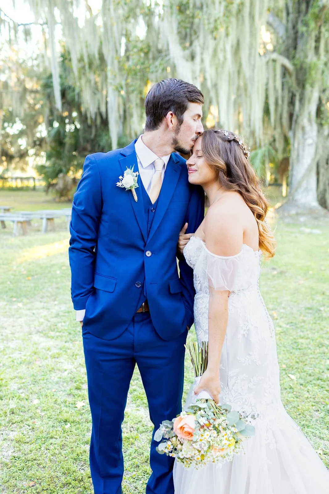 A groom in a blue suit kisses a bride in a white wedding dress during an outdoor wedding ceremony in a park with trees and moss in the background.
