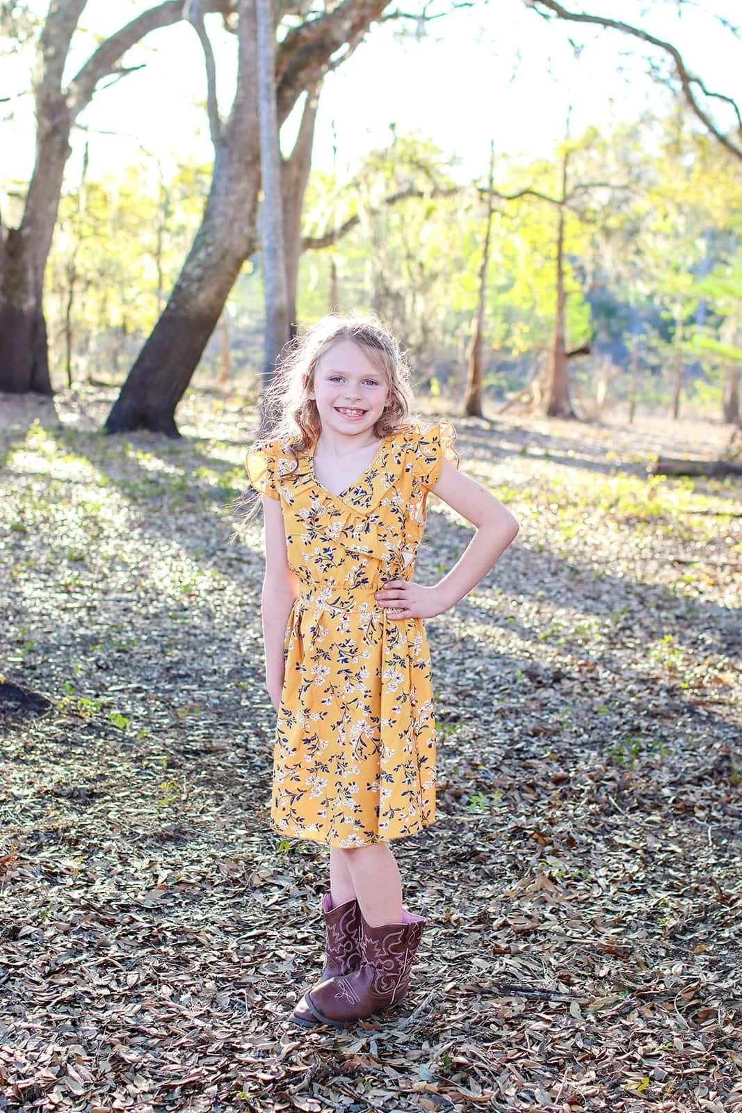 A young girl with curly blonde hair wearing a yellow floral dress and cowboy boots standing in a wooded area with sunlight filtering through the trees.