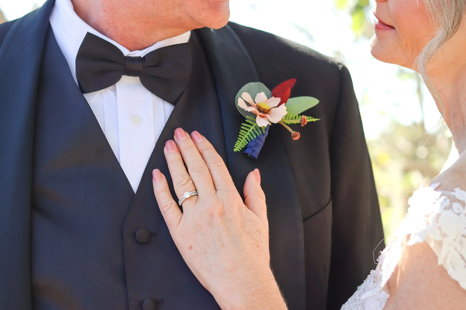 Close-up of a bride touching the lapel of a groom's tuxedo, which has a colorful boutonniere, during a wedding ceremony.