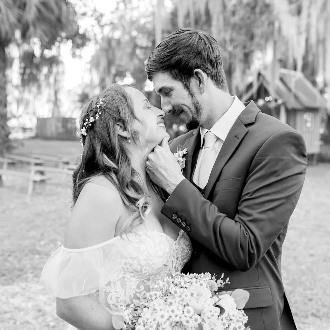 A black and white photo of a couple at their wedding, smiling and touching foreheads outdoors with trees and a wooden building in the background.