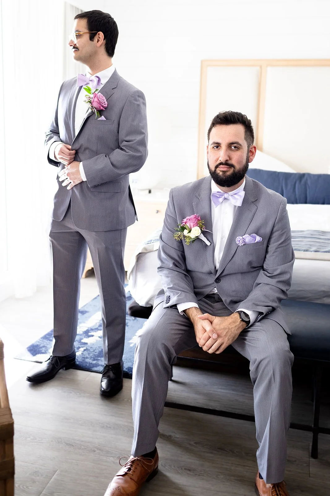 Two men dressed in gray suits with lavender bow ties and boutonnières, one sitting on the bed and the other standing with a hand on his chest, in a bright room with a bed and nightstand.