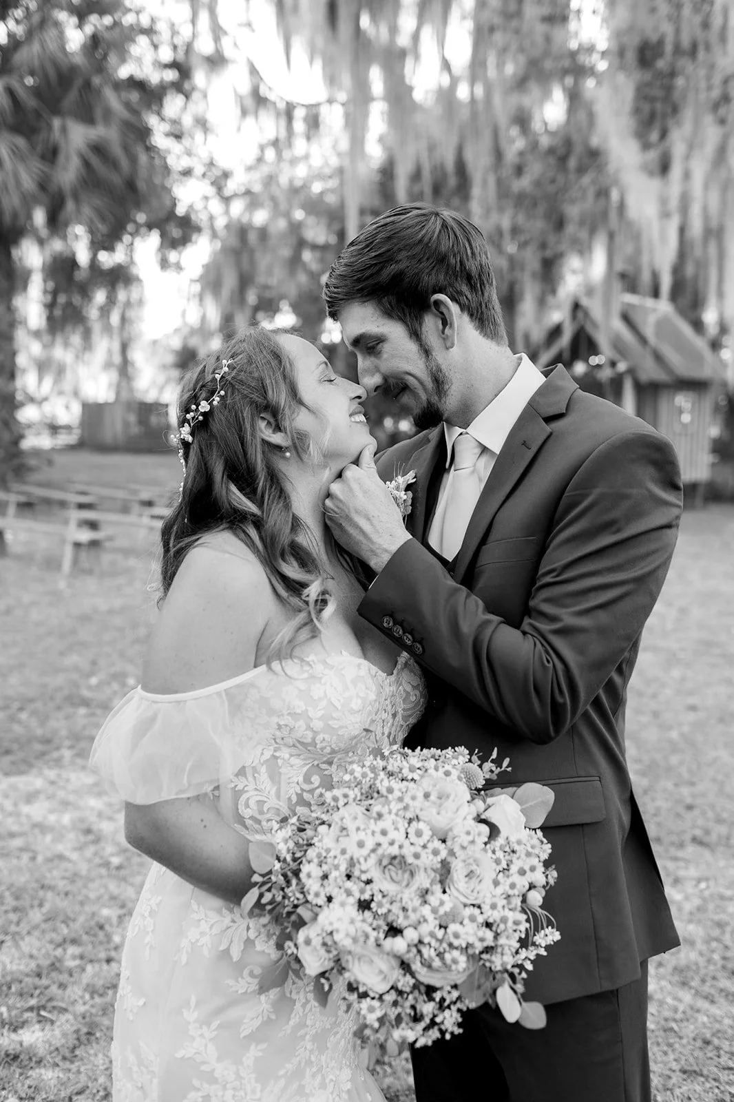 A black and white photo of a bride and groom on their wedding day. The bride holds a bouquet of flowers and is smiling, while the groom gently touches her chin and smiles. They are outdoors with trees and a rustic setting in the background.