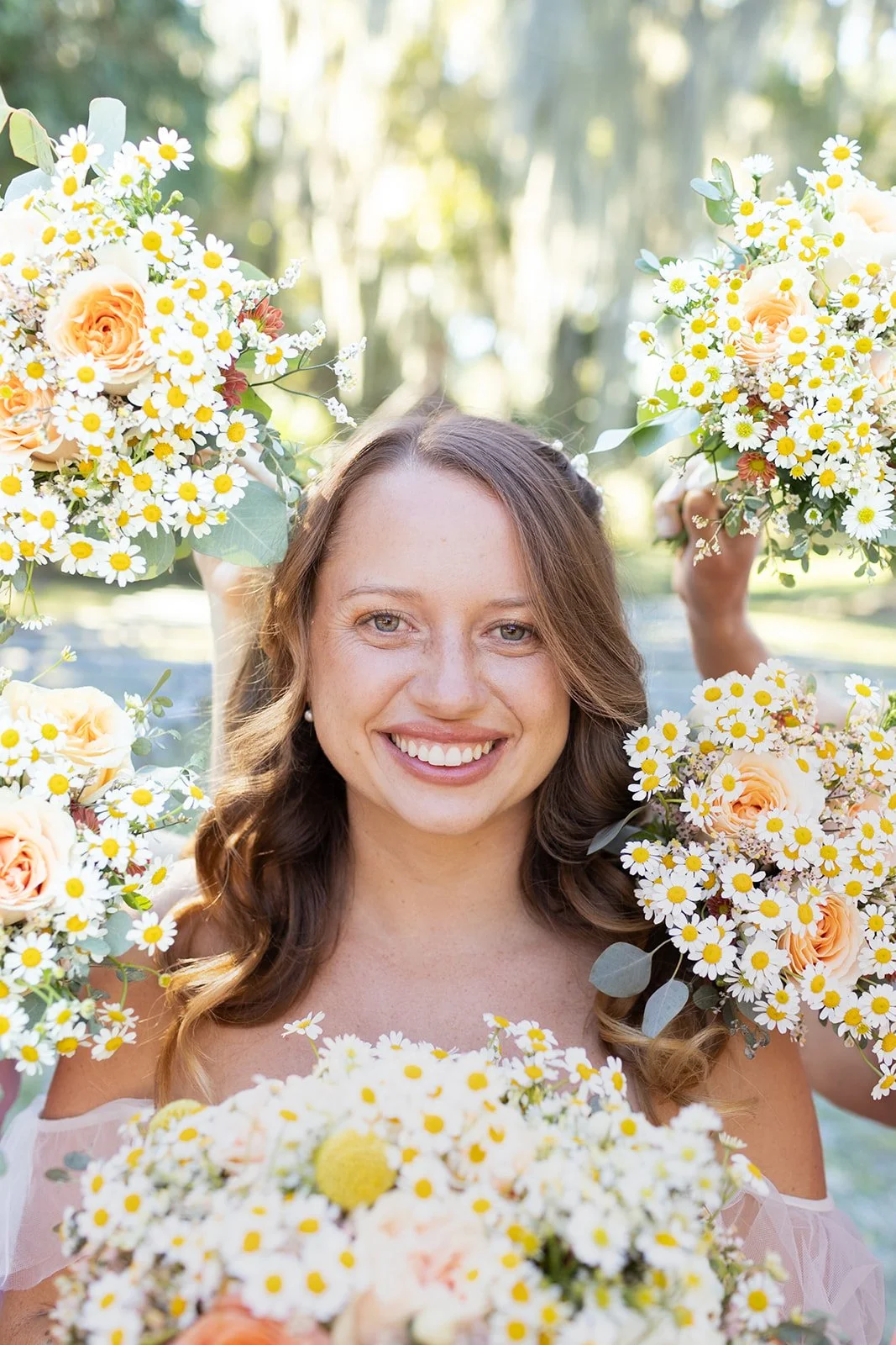 A woman smiling and holding bouquets of flowers, surrounded by more flowers, outdoors in a sunlight setting.