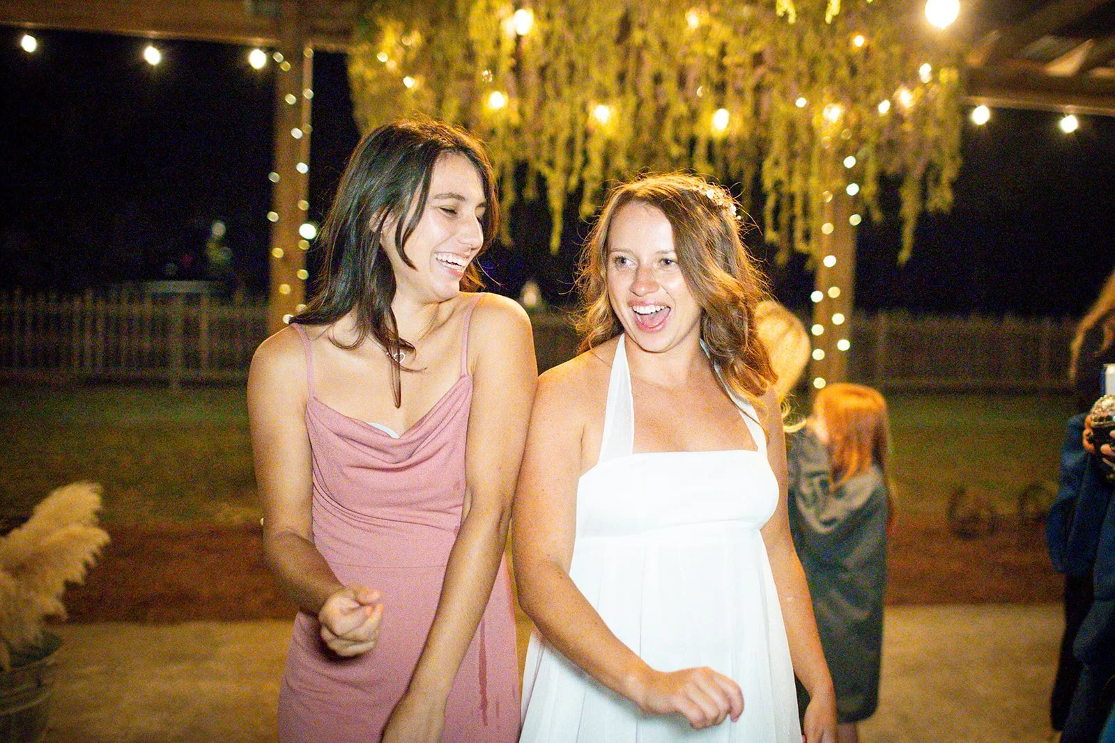 Two women smiling and laughing at an outdoor evening event decorated with string lights and hanging greenery.