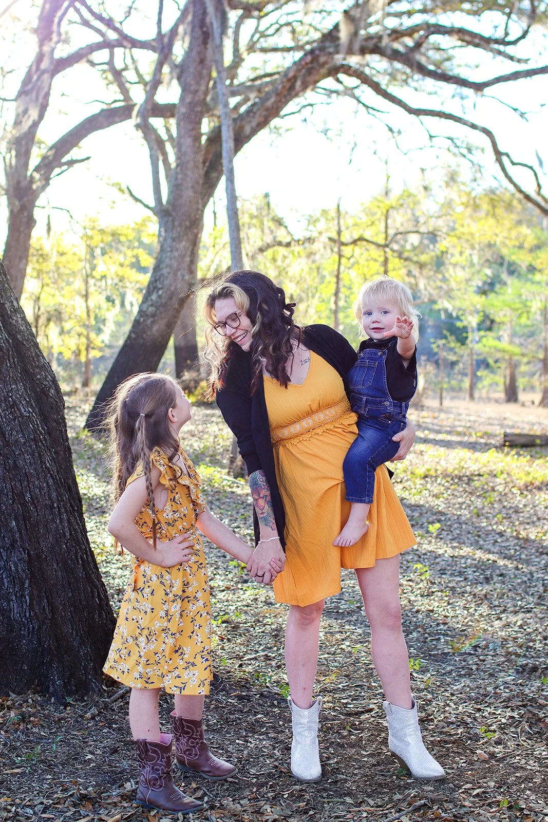 A woman in a yellow dress and black cardigan holding a young boy with blond hair and a dark shirt and overalls, while a girl in a yellow dress and cowboy boots holds her hand. They are outdoors under trees with sunlight streaming through the branches