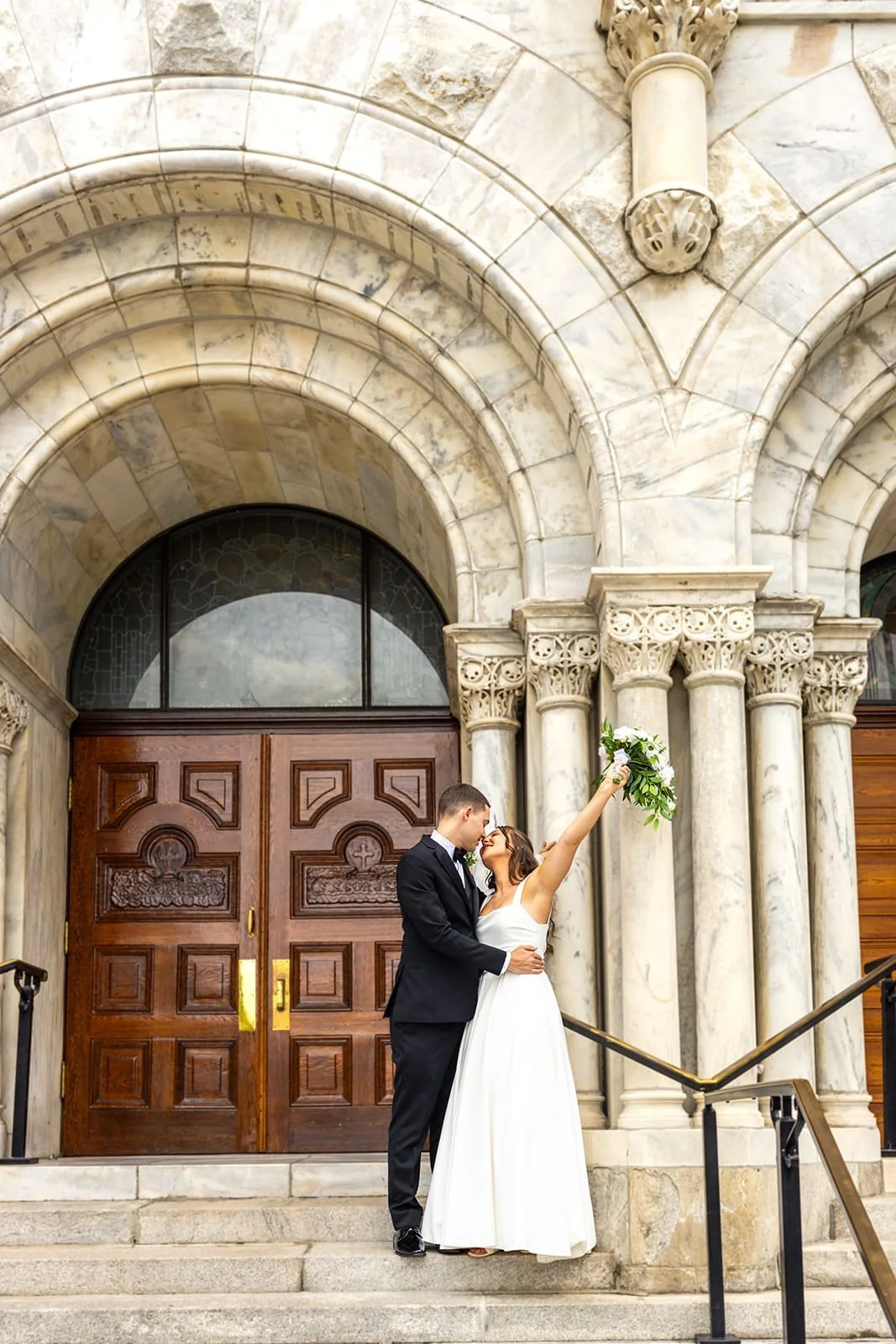 A bride and groom standing on steps outside a church, embracing and smiling at each other, with the bride holding a bouquet of flowers up in the air.