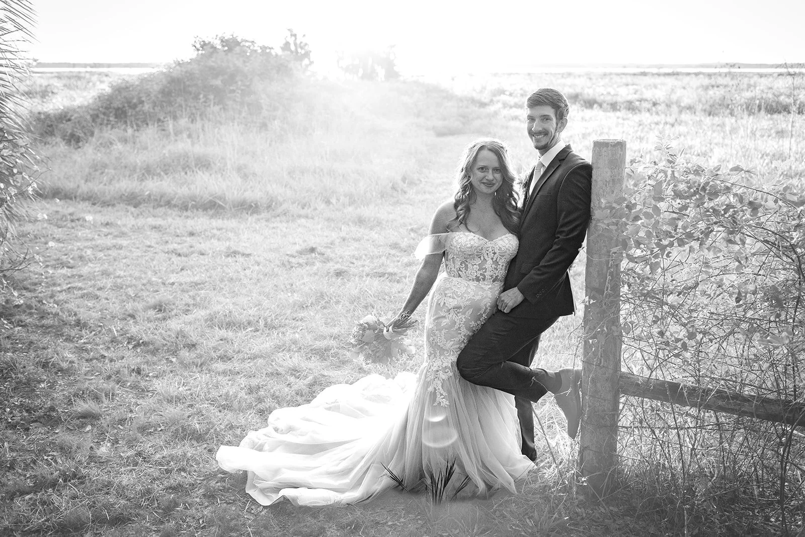 Black and white photo of a bride and groom outdoors. The bride wears a lace wedding gown with off-the-shoulder sleeves, holding a bouquet. The groom in a suit leans against a wooden post, both smiling.