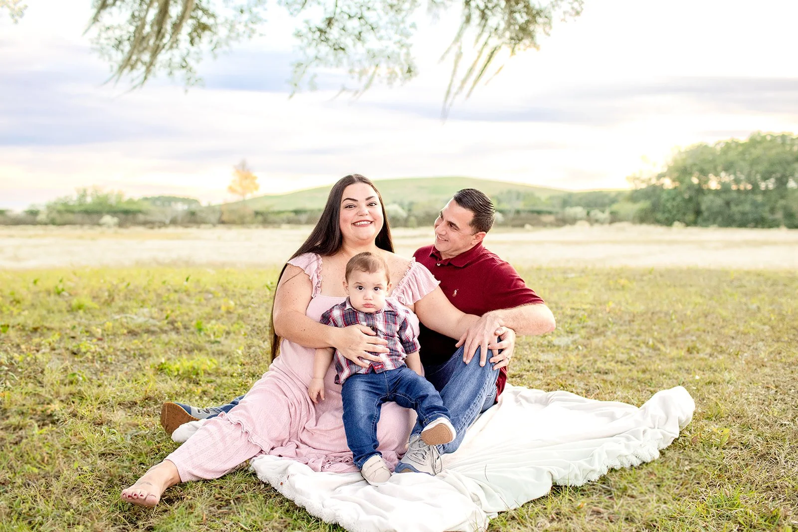 A family of three sitting on a white blanket on the grass in a park. The woman is wearing a pink dress, the man a red shirt, and the child a plaid shirt. The woman is holding the child, and the man is sitting close to her. They are smiling at each ot