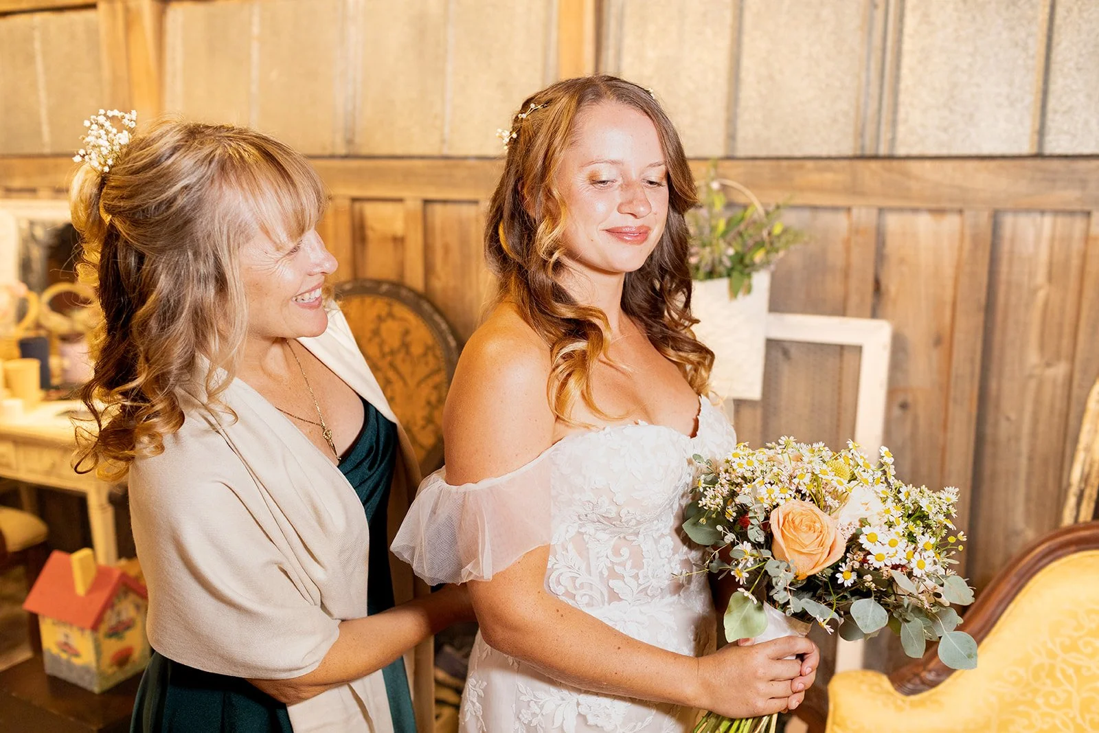 A bride holding a bouquet of flowers, standing with a woman at a wedding ceremony in a rustic indoor setting.