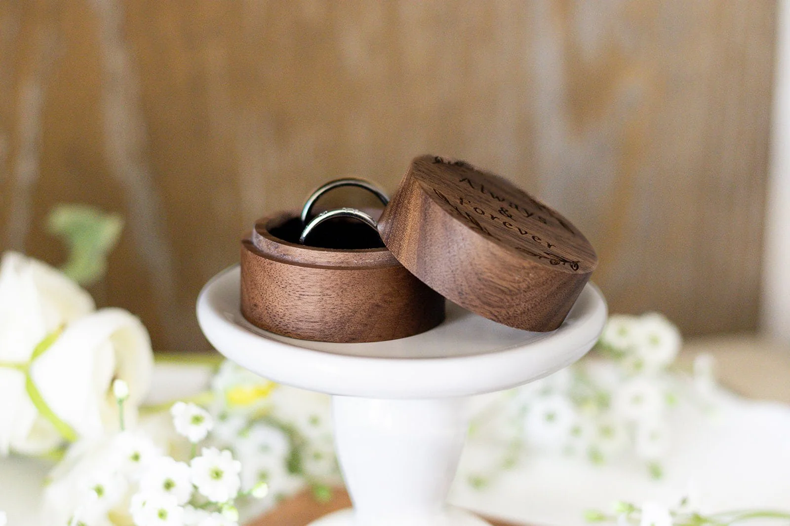 Wooden ring box open with two wedding bands inside, placed on a white stand. Flowers are blurred in the background.