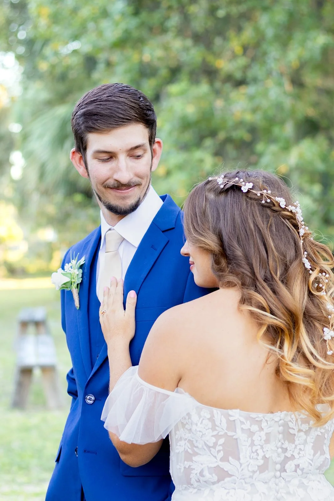 A couple on their wedding day outdoors, the woman wearing a lace off-shoulder dress with a floral headpiece, touching the man's chest, who is dressed in a blue suit with a boutonniere.