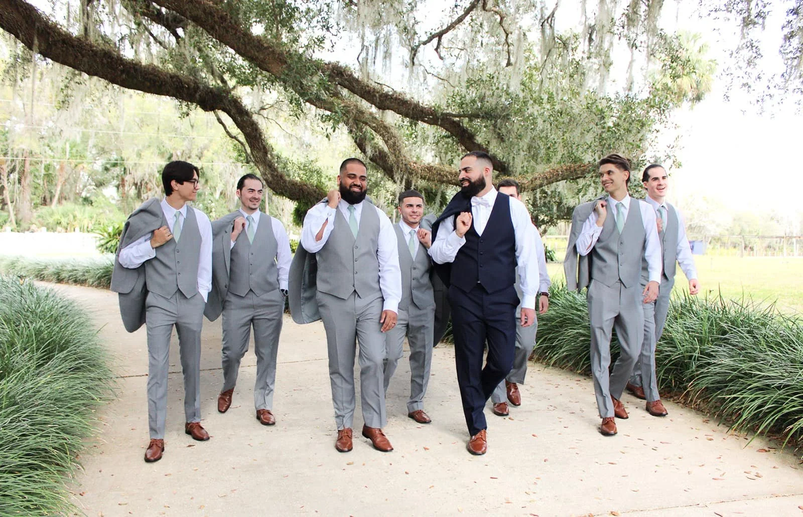 A group of seven men in suits walking outdoors under a large tree, smiling and talking, with some carrying their suits over their shoulders.