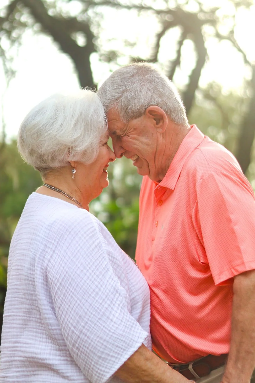 An elderly couple has their foreheads touching, smiling and demonstrating affection outdoors during daytime.