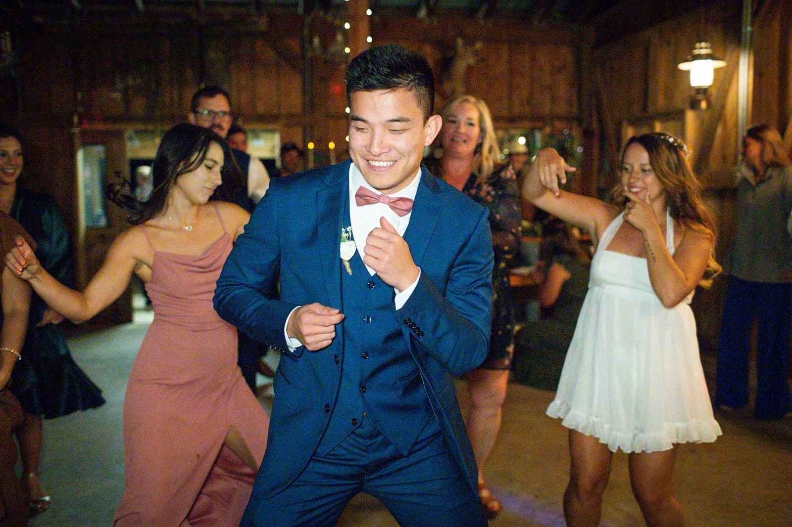 A young man in a blue suit and pink bow tie dancing with a broad smile at a party, surrounded by women also dancing in a warmly lit wooden venue.