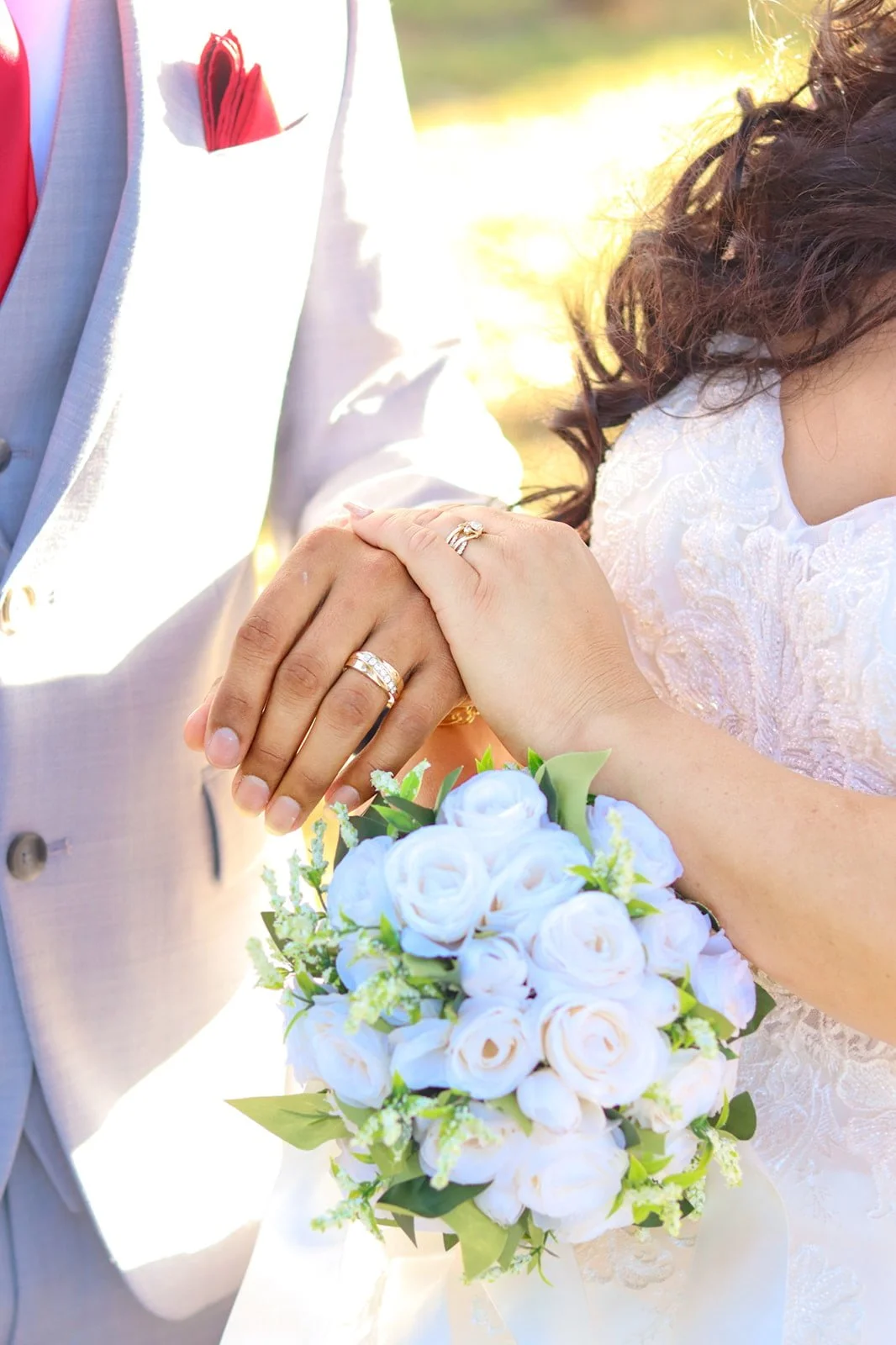 Close-up of newlywed couple holding hands, showing wedding rings, with bride holding a white bouquet of roses. The groom is wearing a light-colored suit with a red pocket square. The bride is wearing a lace wedding dress. The background is bright and