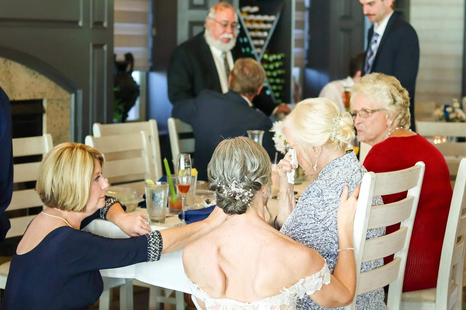 Group of women at a wedding reception, with one elderly woman wiping tears and others comforting her, seated around a table with drinks.