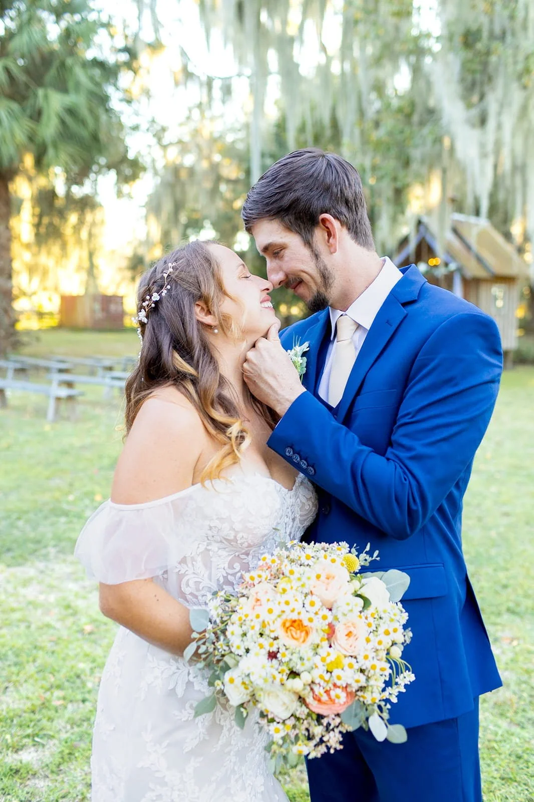 A bride and groom sharing an intimate moment outdoors, with trees and a rustic building in the background. The bride holds a large bouquet of flowers, and both are smiling as they touch foreheads.