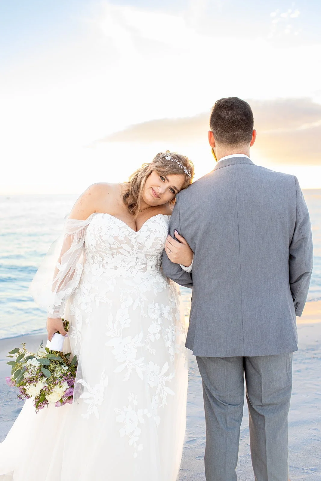 Bride in a white wedding dress with floral embroidery leaning her head on groom's shoulder at the beach during sunset.