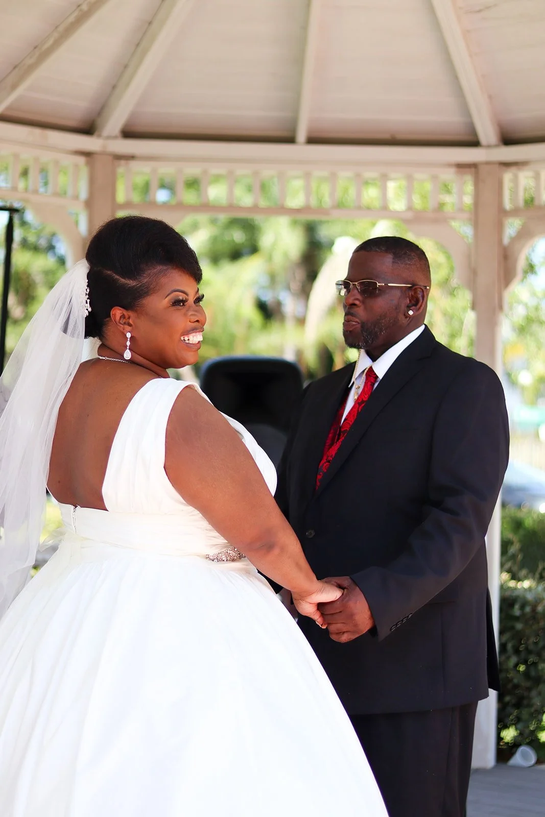 Happy couple at a wedding ceremony holding hands under a gazebo, with the bride in a white wedding gown and the groom in a black suit with a red tie, surrounded by outdoor greenery.