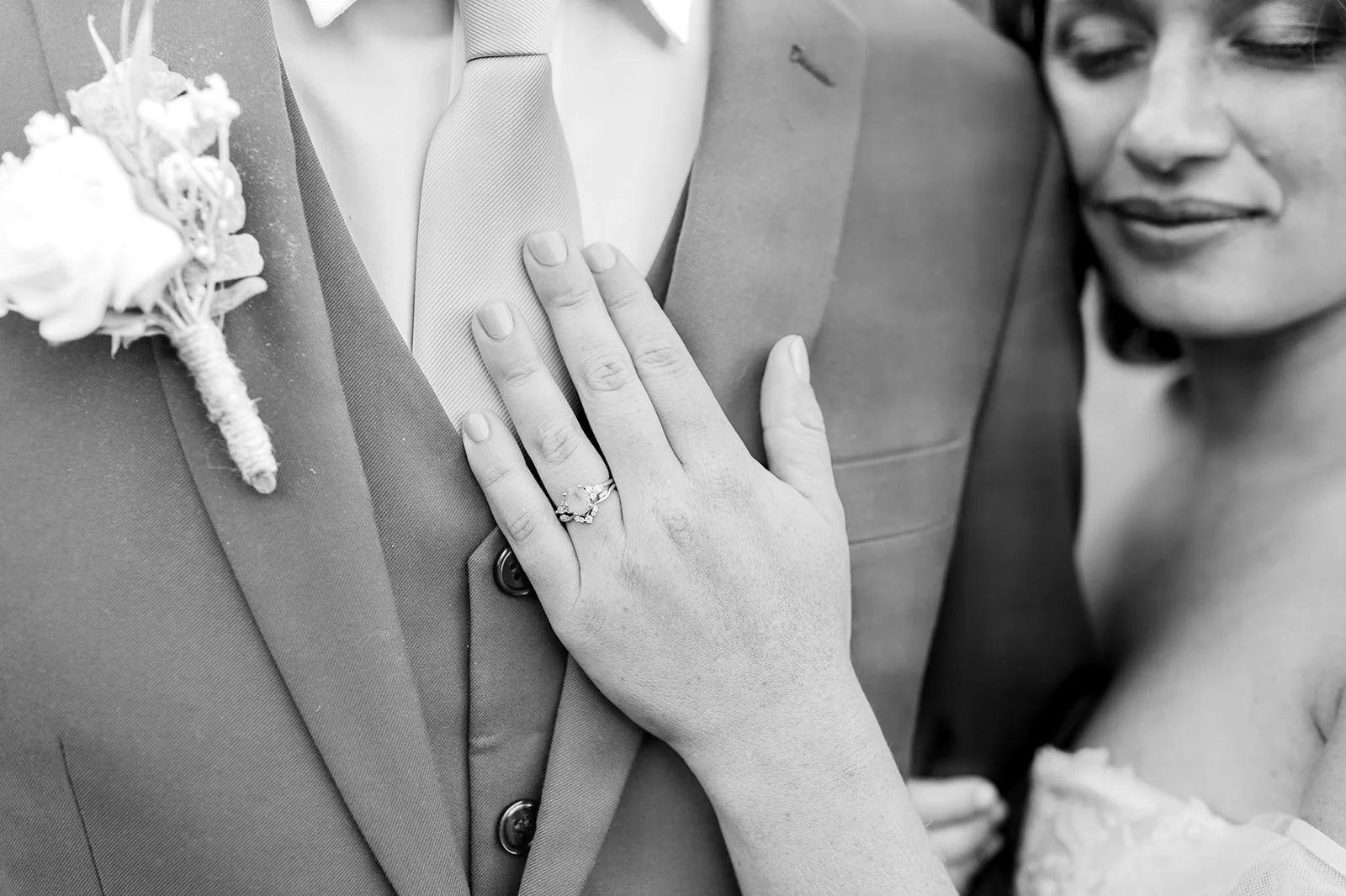 Close-up of a bride's hand with a wedding ring resting on the chest of a groom wearing a suit with a boutonniere, on a wedding day. The bride's face is slightly visible, smiling in the background.