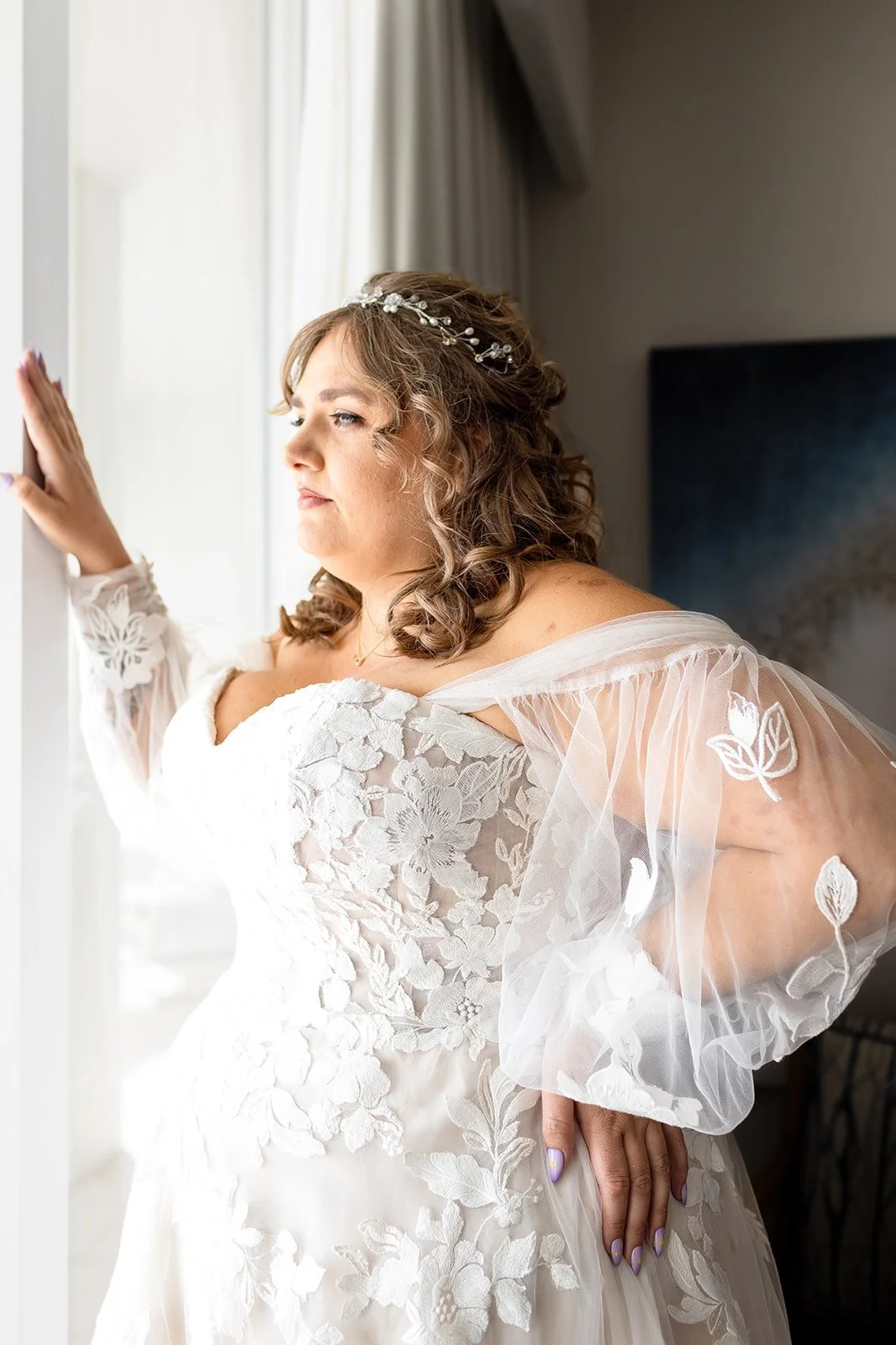 A bride in a white lace wedding dress with sheer floral embroidered sleeves looking out a window with her hand on the window frame.