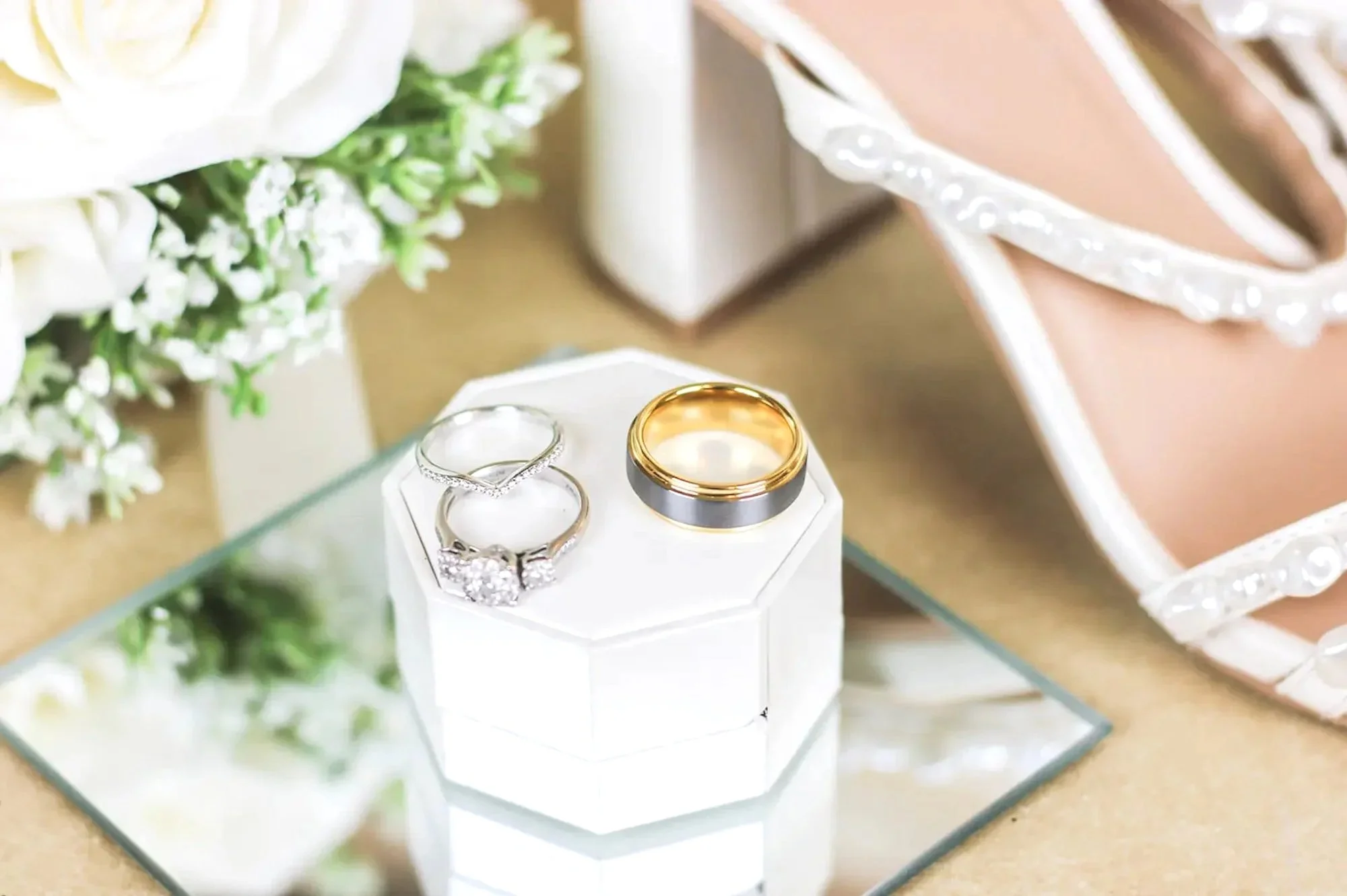 classy wedding bands laid on a ring box, evergreen florals in back next to brides white heel laced with pearls