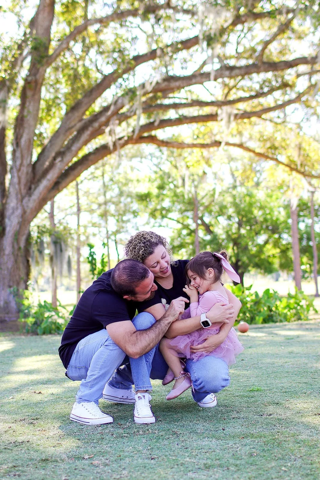 Family of three, including a man, woman, and girl, laughing and playing outdoors on a grassy yard under a large tree on a sunny day.
