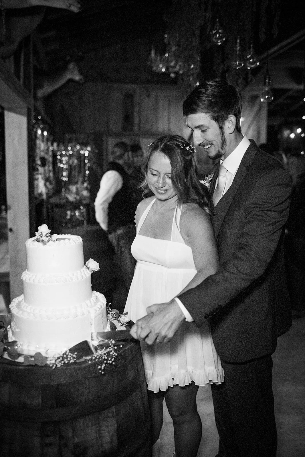 Black and white photo of a wedding reception where a bride and groom are cutting a wedding cake together. The bride wears a sleeveless dress with spaghetti straps and has a small floral hair accessory. The groom wears a dark suit with a tie. They are