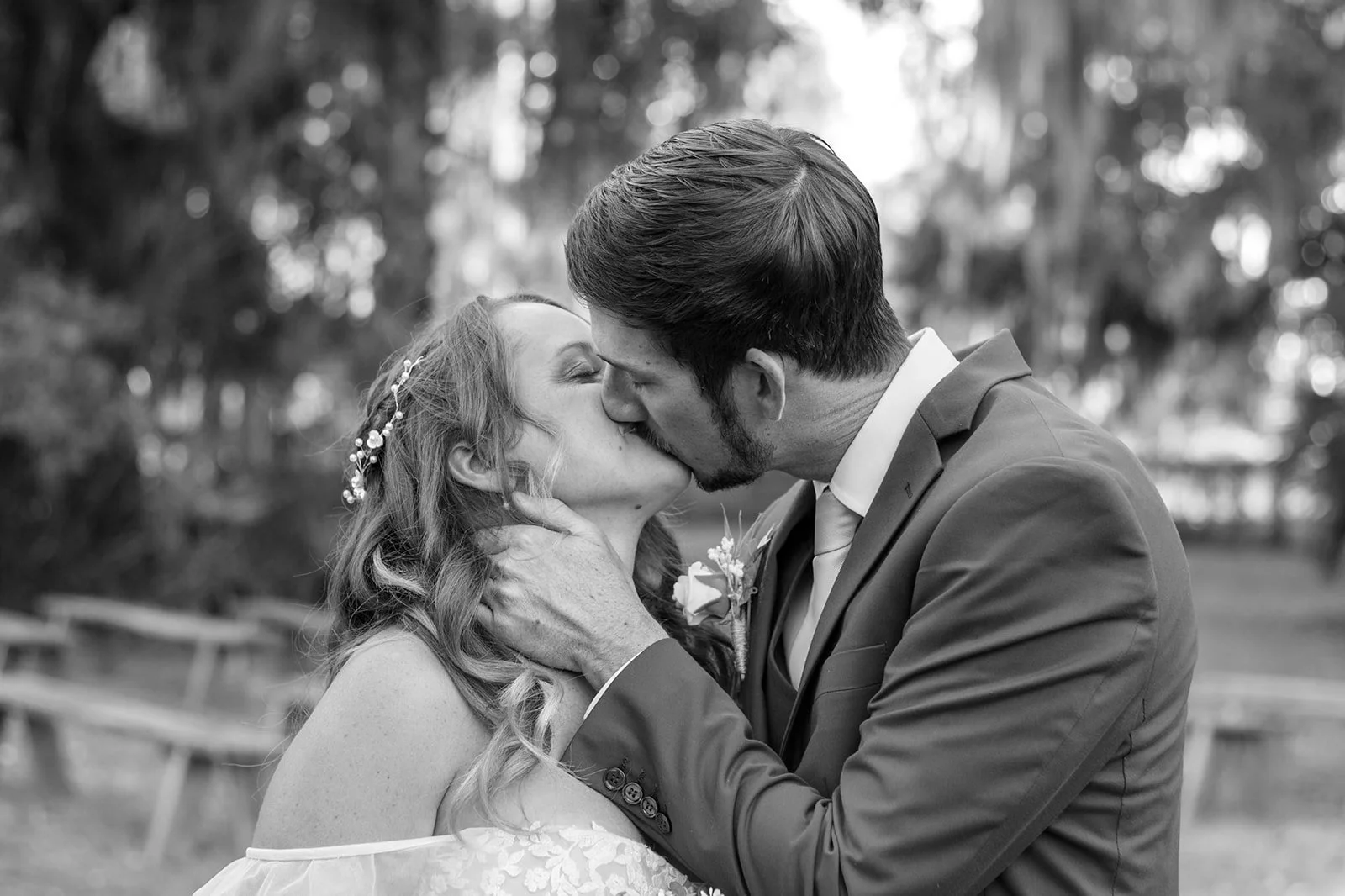 A black and white photograph of a couple kissing outdoors. The woman has long wavy hair with a decorative headband, and the man is wearing a suit with a tie. They are embracing each other amidst blurred trees in the background.