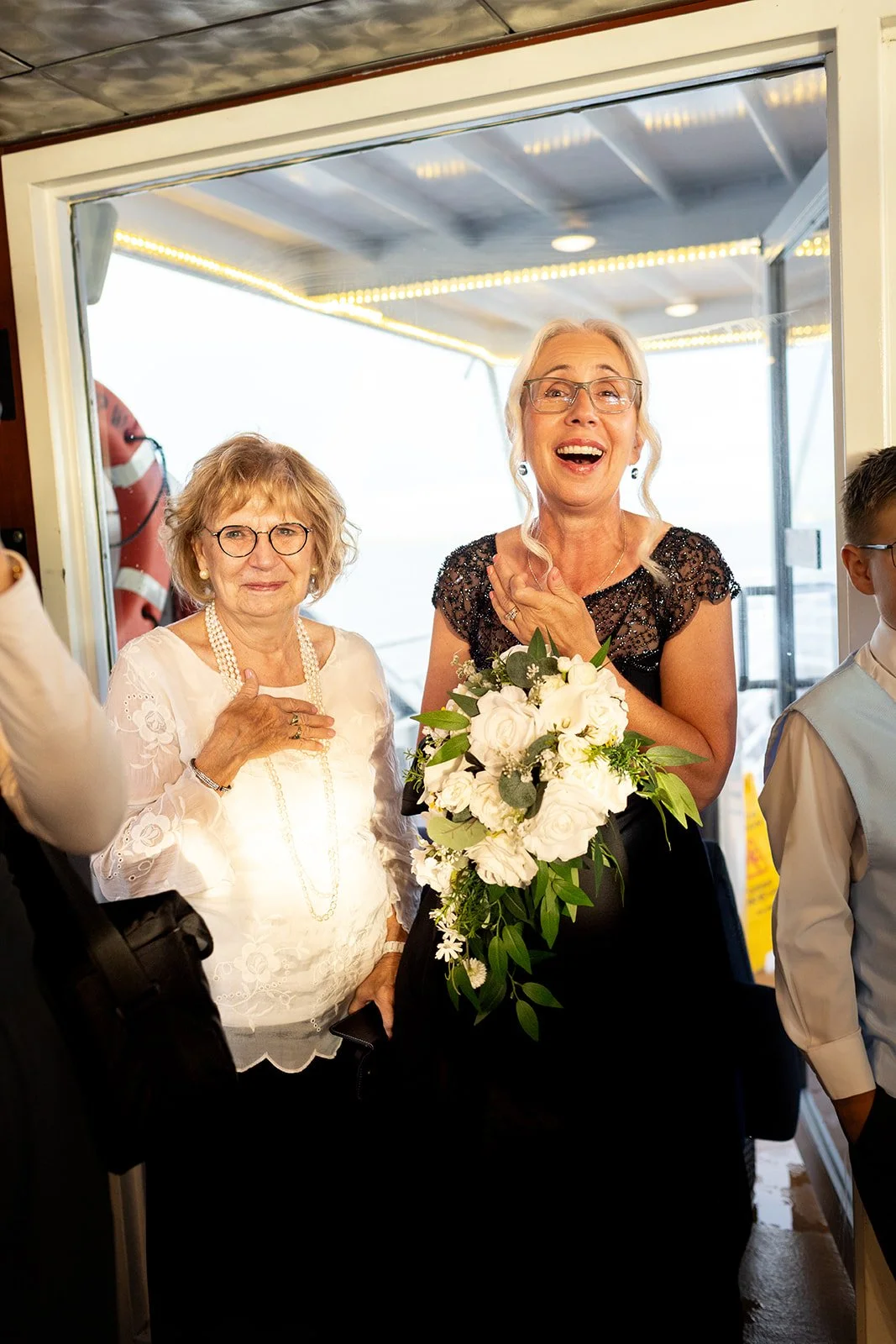 Two women at a celebration, one holding a bouquet of white flowers, the other with her hand on her chest, smiling and appearing joyful.