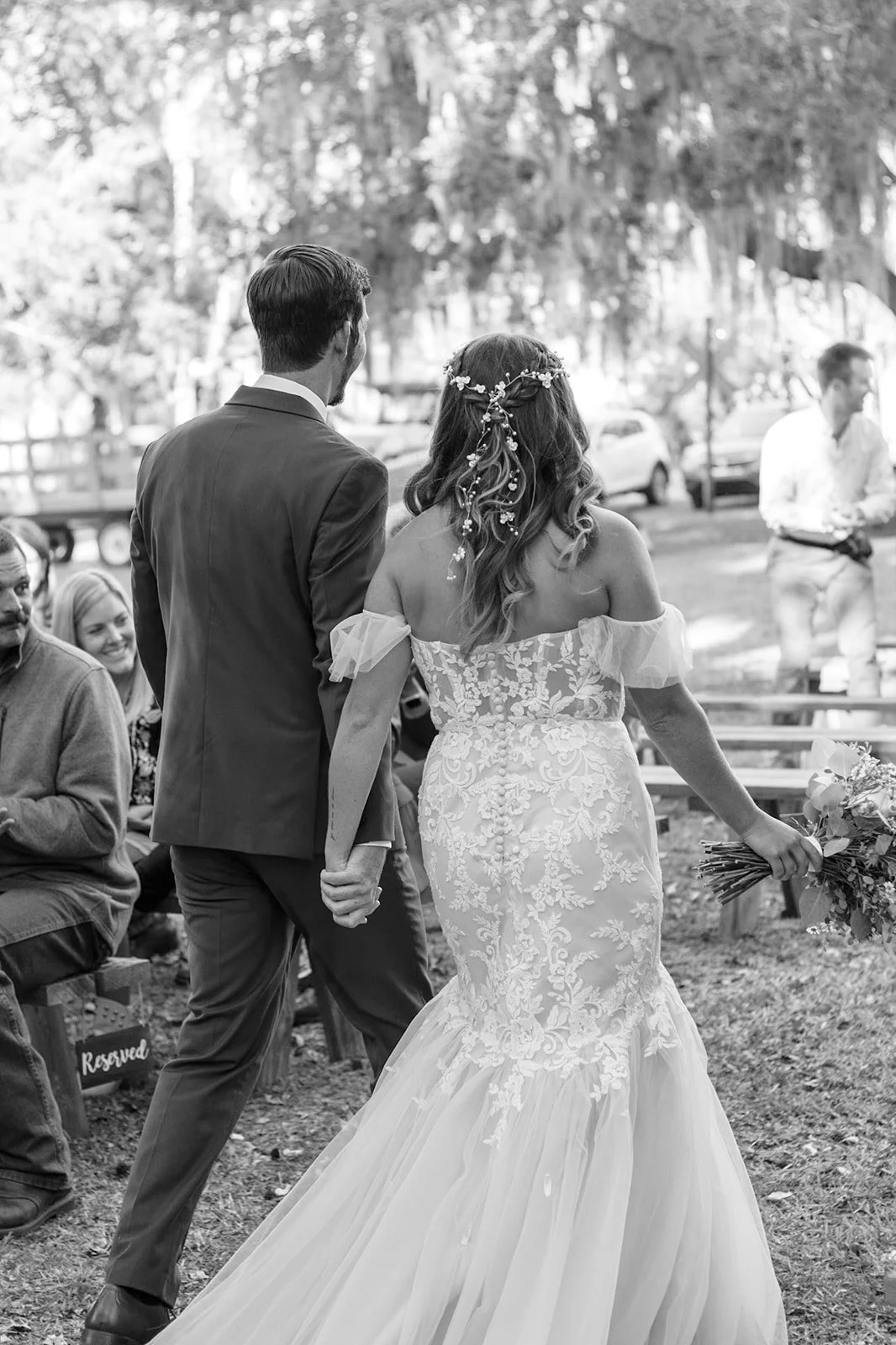 A bride and groom holding hands walking at an outdoor wedding ceremony, with guests seated and trees in the background.