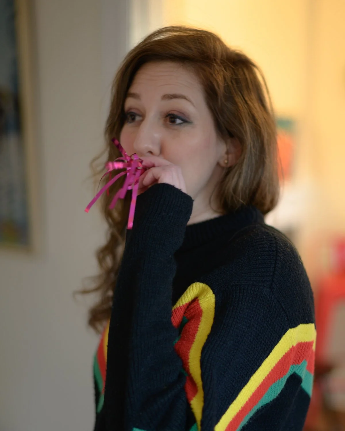 A woman with wavy brown hair wearing a black sweater with colorful rainbow stripes, with a party blower noisemaker in her mouth standing in a doorway.