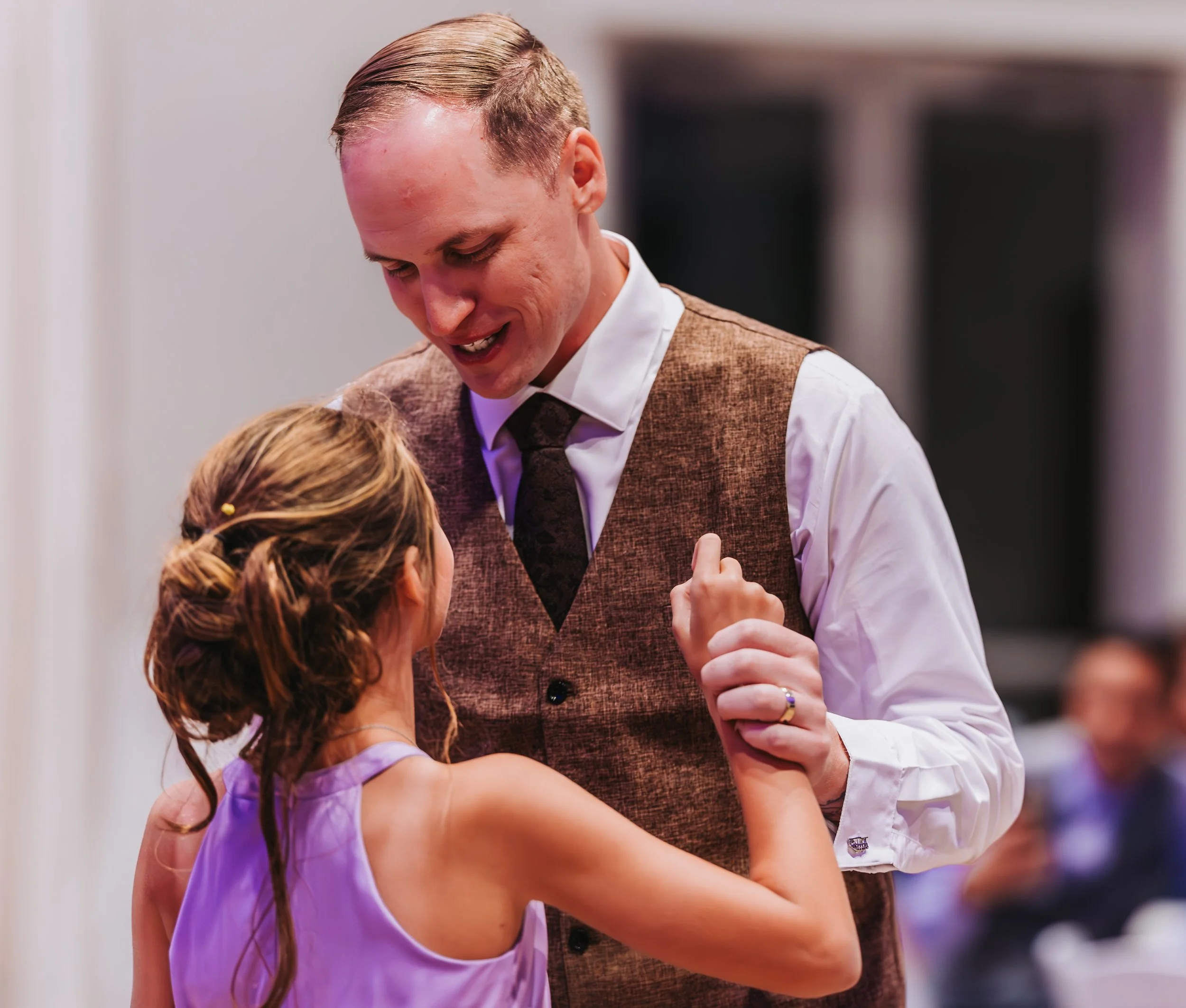 A man and a young girl dancing together at an indoor event; the man is smiling and holding the girl's hand, wearing a vest and tie, while the girl has her hair styled and wears a purple dress. western wedding photographer az rustic wedding photograph