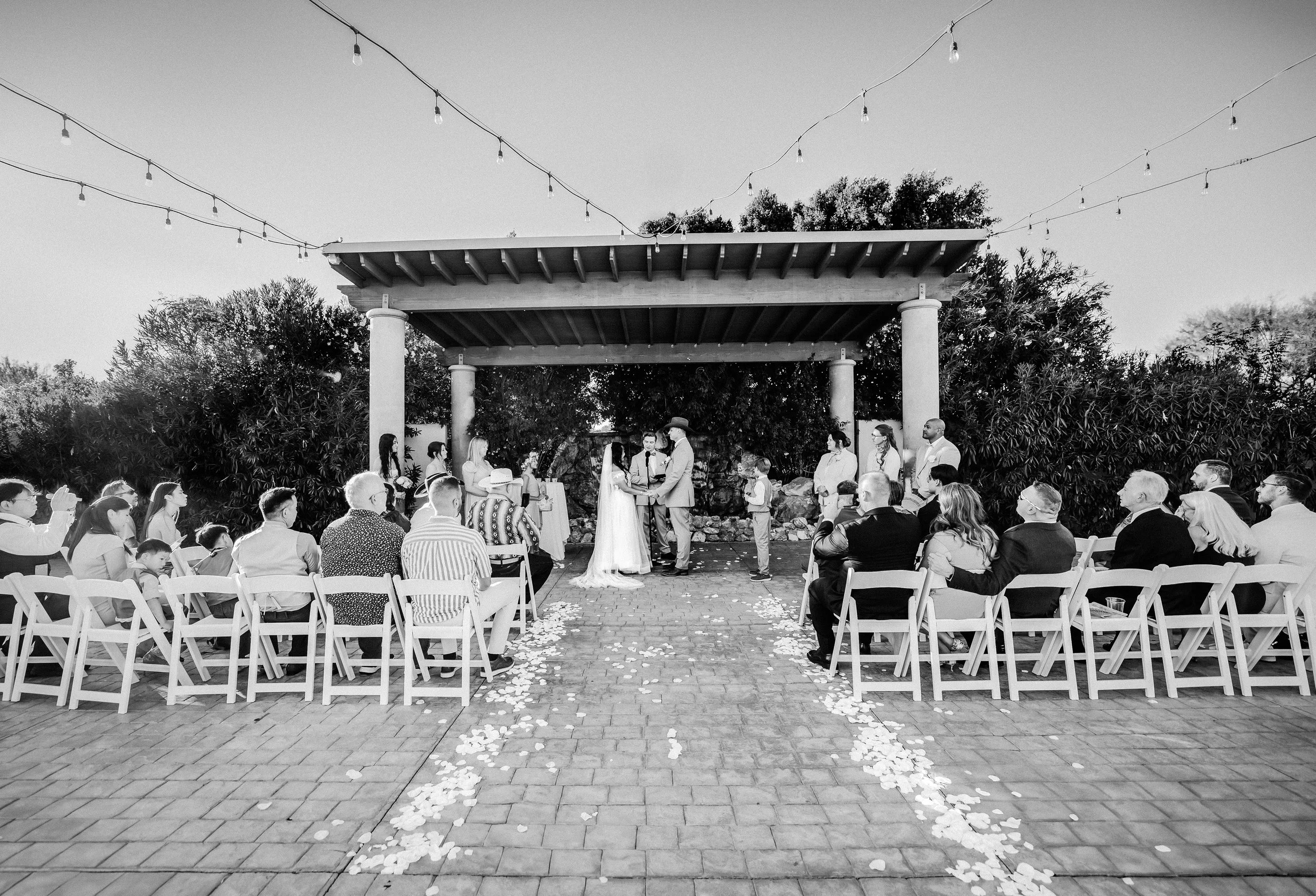 Black and white photo of an outdoor wedding ceremony with guests seated on white chairs, a bride and groom exchanging vows under a pavilion, surrounded by trees and string lights. affordable wedding photographer phoenix az
desert wedding photographer