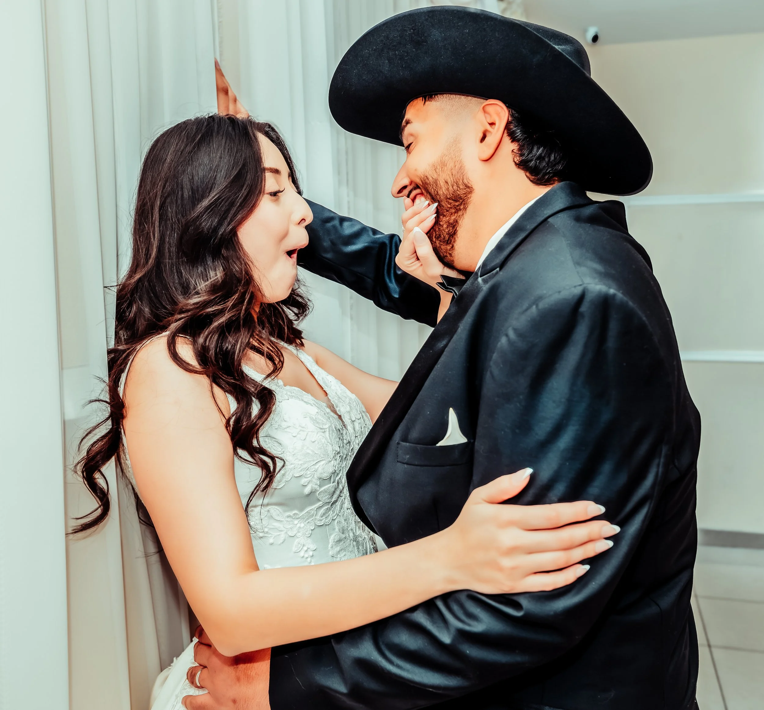 A couple during their wedding, with the bride in a white lace dress and the groom in a black tuxedo and cowboy hat, sharing a joyful moment indoors.indoor Mexican wedding photographer phoenix az