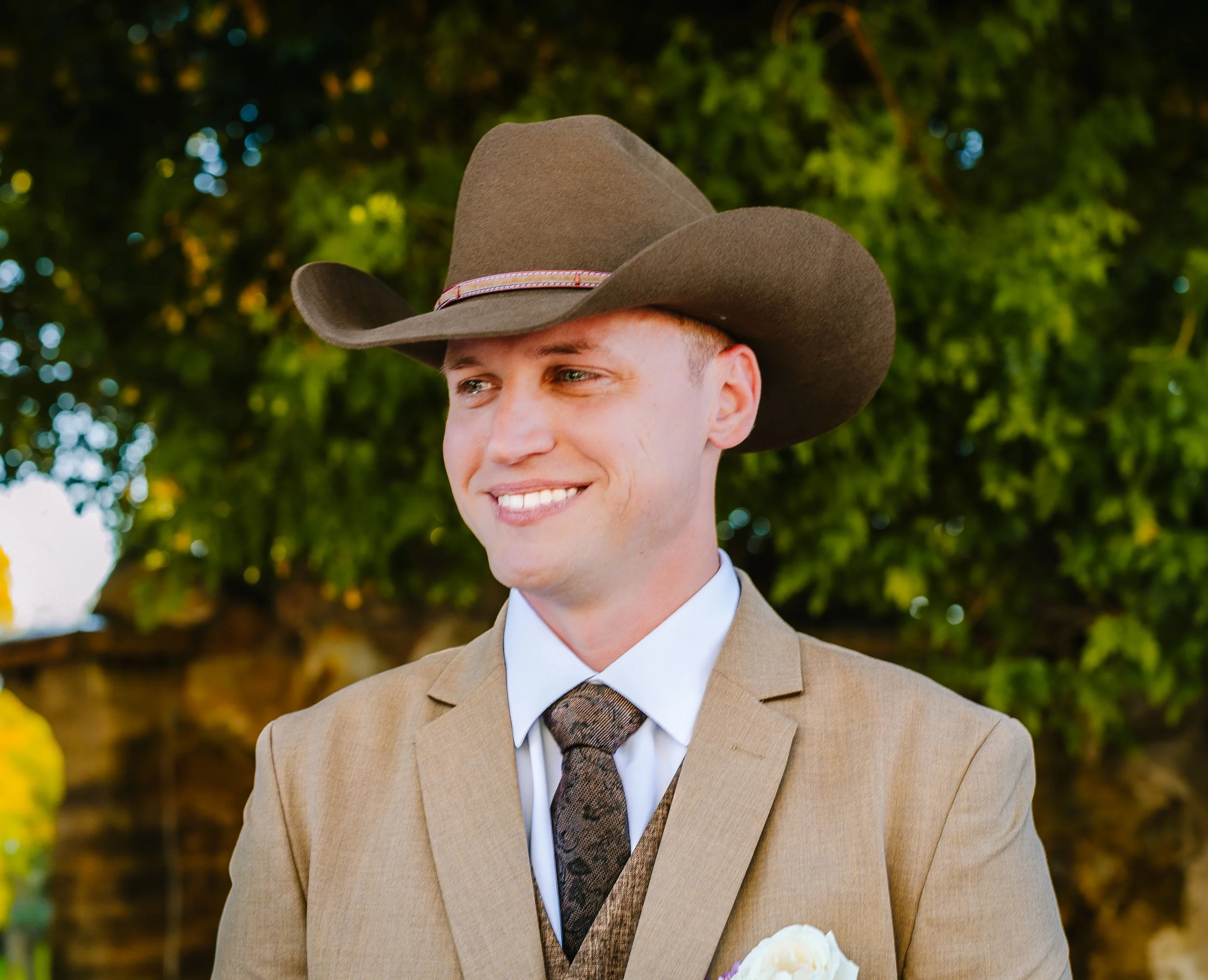 A smiling man wearing a tan suit, white dress shirt, patterned tie, and a wide-brimmed cowboy hat, standing outdoors with green foliage in the background. traveling wedding photographer az wedding photographer Prescott az wedding photographer flagsta