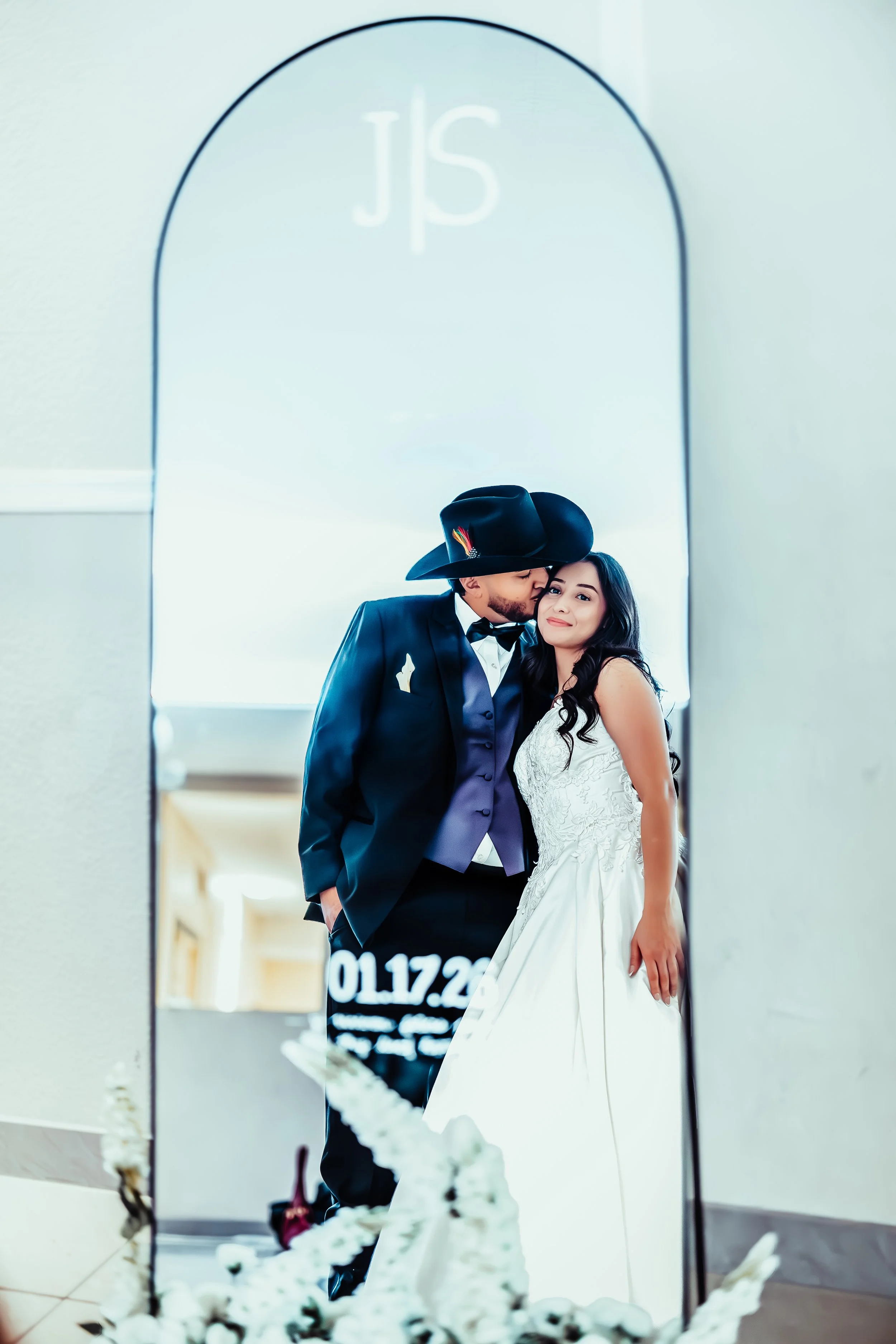 A couple in wedding attire, with the groom in a suit and hat and the bride in a white wedding dress, seen through a decorative mirror. indoor Mexican wedding photographer phoenix az